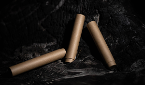 Three cylindrical, eco-friendly cannabis packaging tubes made from brown, biodegradable material, placed on a dark, textured surface. The tubes have a minimalist design, emphasizing sustainability and innovative closure solutions.