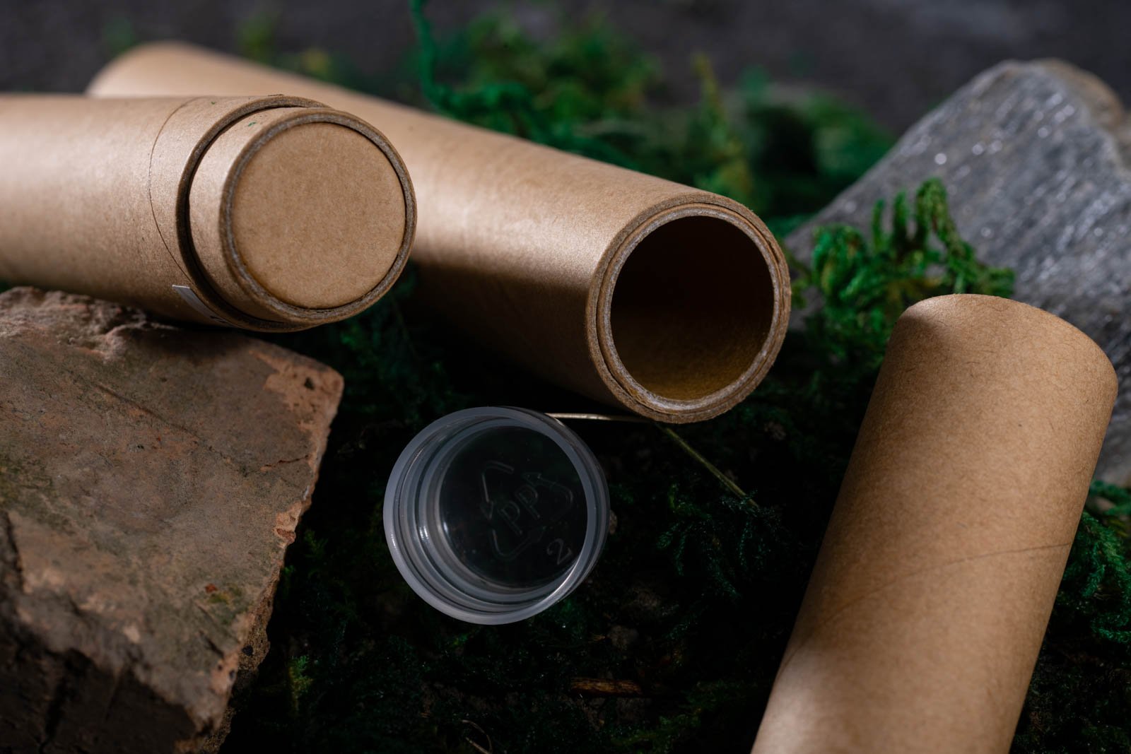 A close-up of eco-friendly cylindrical cannabis packaging tubes made from biodegradable brown material, placed on a natural surface with moss, rocks, and wood. One tube is open, revealing a plastic inner liner with a recycling symbol, highlighting a combination of sustainability and secure product storage.